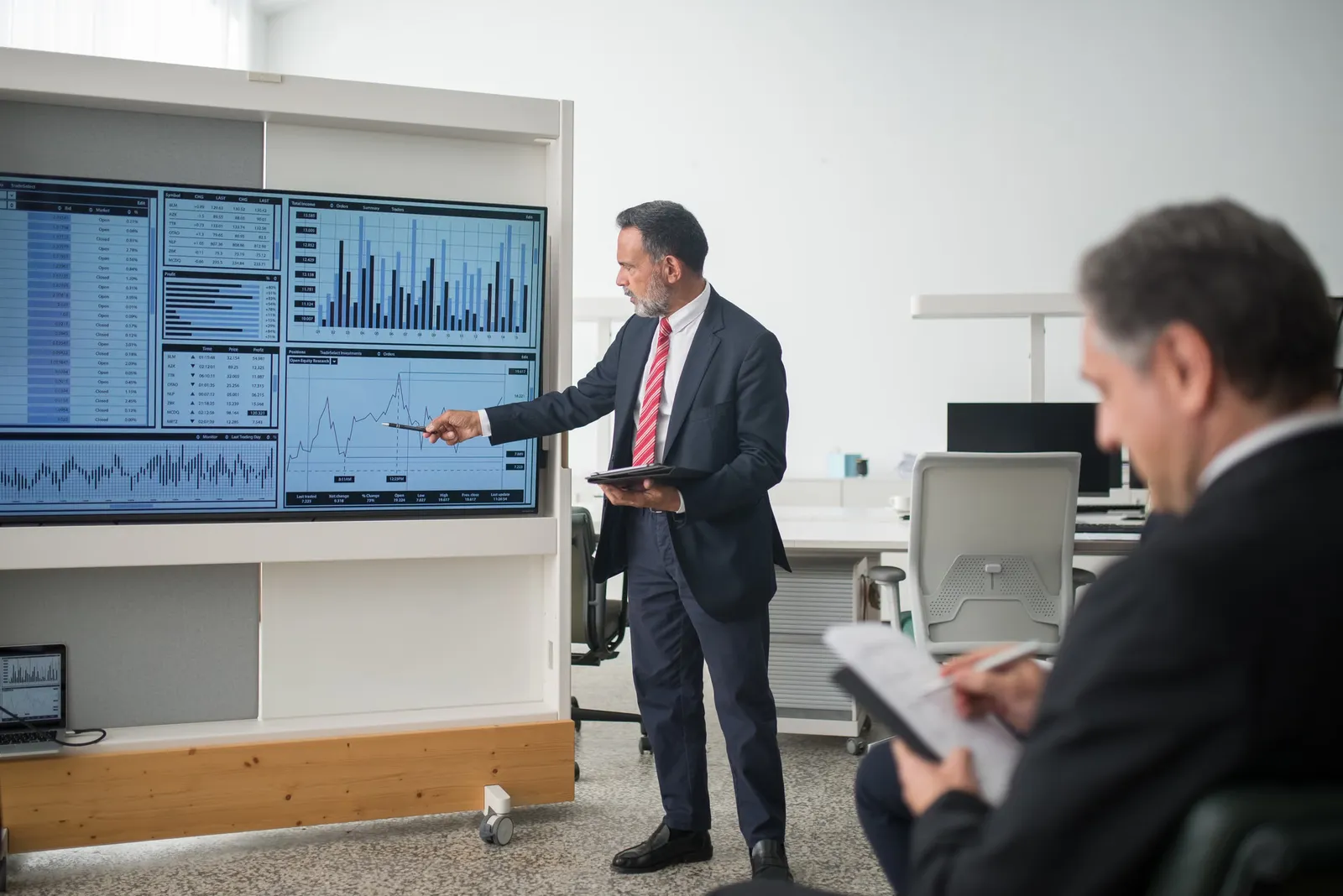 A man presenting program performance charts on a large screen while a colleague takes notes